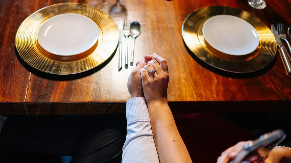 Deux verres et une bougie sur une table basse, la main d'une femme posée près de celle d'un homme, lumière tamisée
