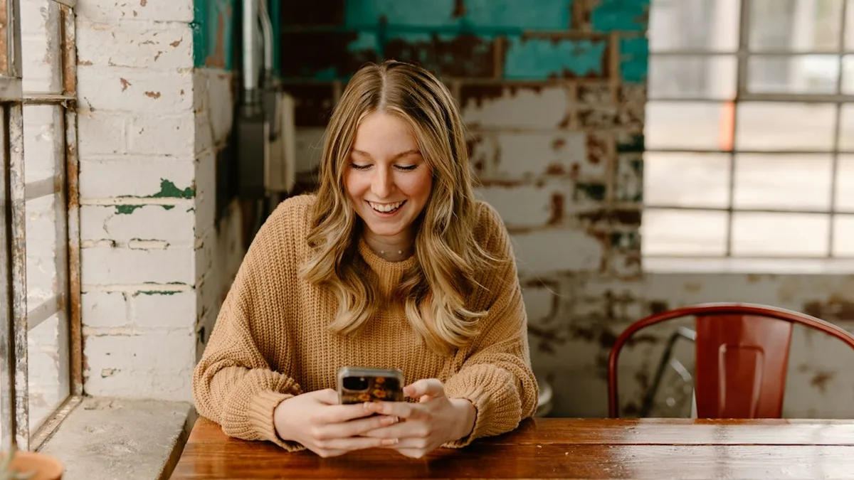 Une femme souriante lit un message sur son téléphone, attablée dans un café aux murs de brique